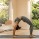 A female yoga instructor, wearing a grey fitted tank top and leggings, performs the advanced Wheel Pose (Urdhva Dhanurasana), creating a deep, symmetrical backbend supported by her hands and feet on a wooden studio floor.